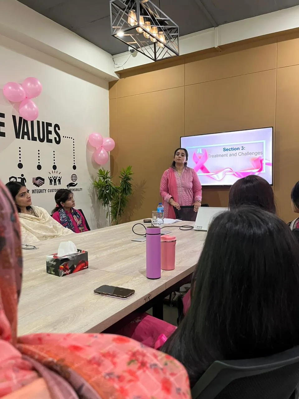 A speaker in pink standing at the head of the WorkBench board room table with a slide on the screen behind her, while women seated along the table listen and pink balloons hang on the Core Values wall.