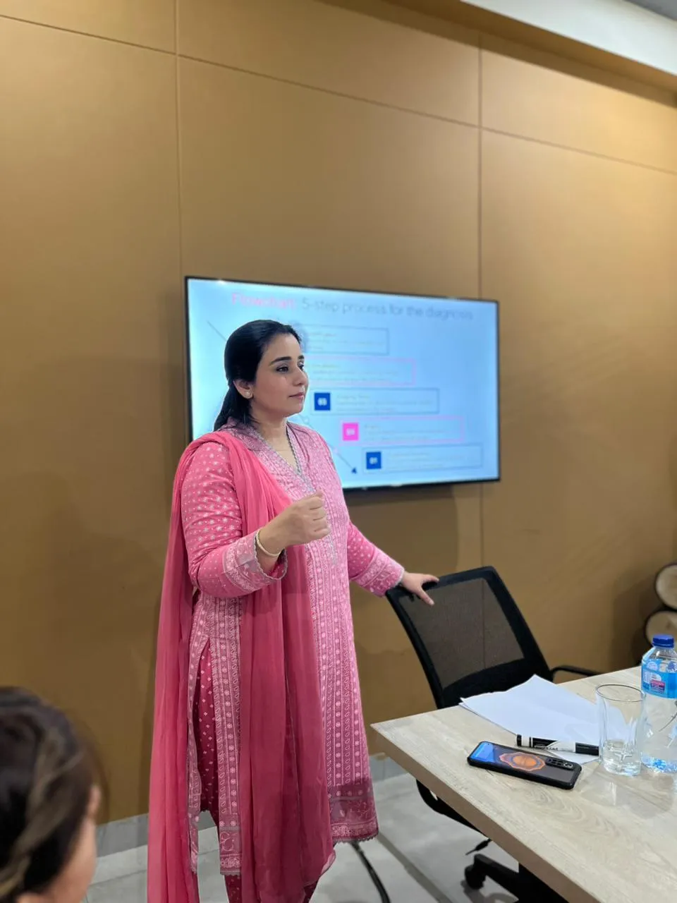 A speaker in a pink shalwar kameez standing beside the meeting room screen at WorkBench, mid sentence, with slides showing behind her.