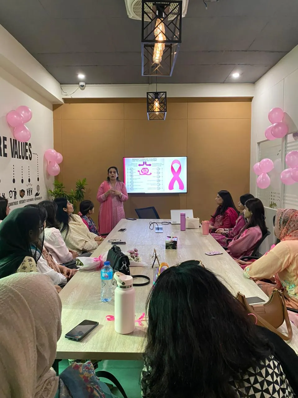 A session in the WorkBench meeting room: a speaker presenting a slide with a large pink ribbon on it, women seated along both sides of the long table, pink balloons taped to the walls.