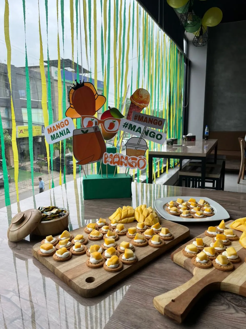 The mango party table at WorkBench seen through hanging green and yellow streamers, with cardboard mango props on sticks and trays of mango canapes on the counter.