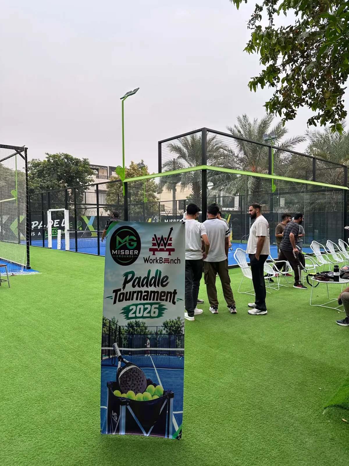 A pull-up banner reading Paddle Tournament 2026, co-branded Misber Grounds and WorkBench, standing on artificial grass beside glass-walled padel courts, with players gathering by a row of white chairs.
