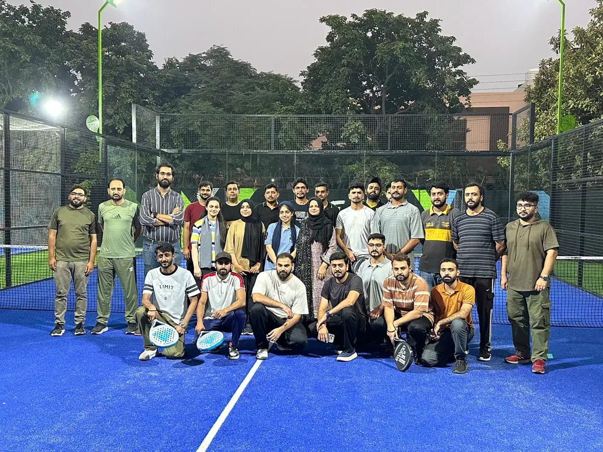 A large group of WorkBench members posing for a group photograph on a blue padel court under floodlights, the front row crouched with padel rackets, a glass and mesh court wall and trees behind them.