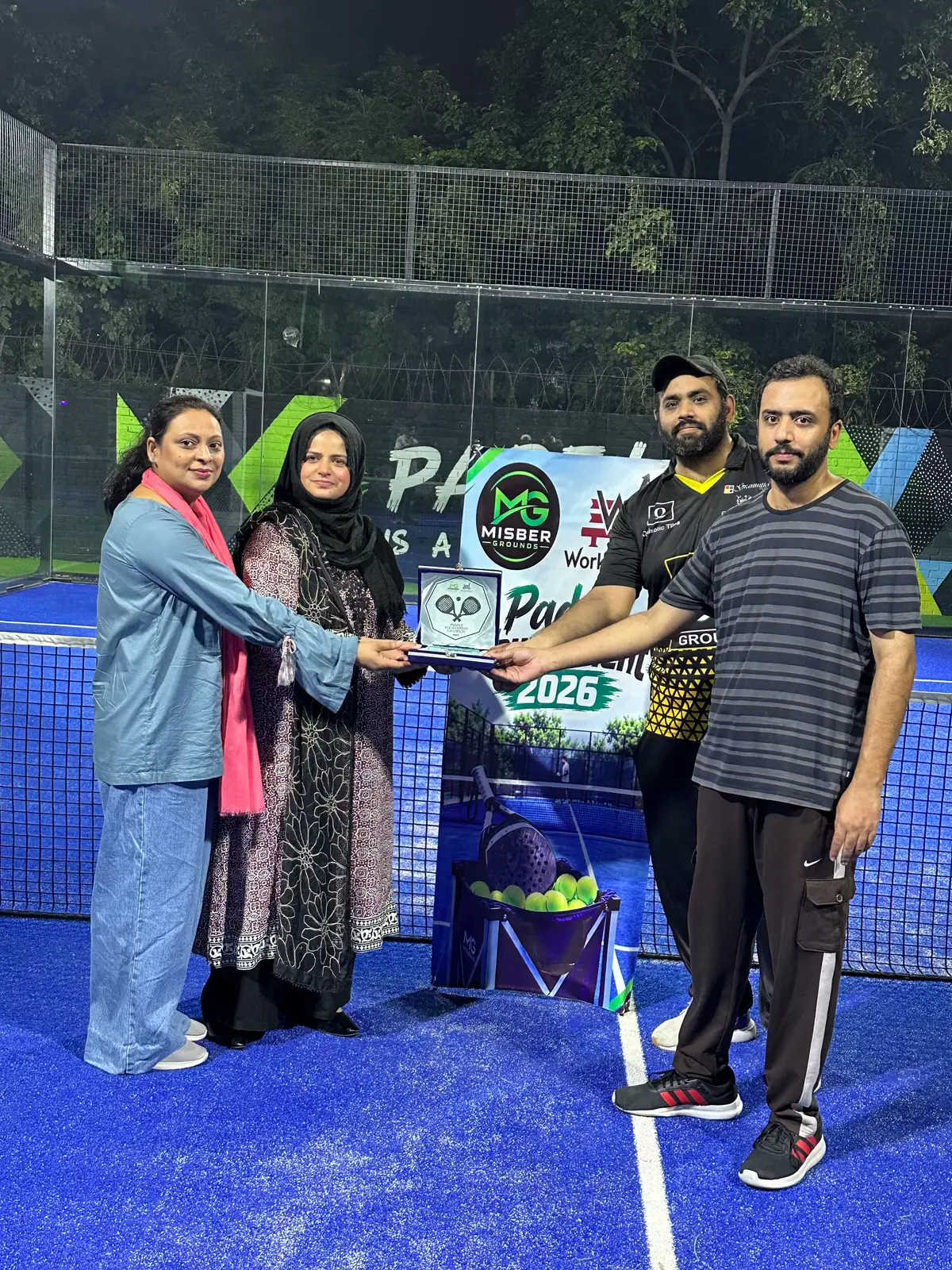Two women presenting a plaque in a blue presentation box to two men on a blue padel court at night, in front of the Paddle Tournament 2026 banner.