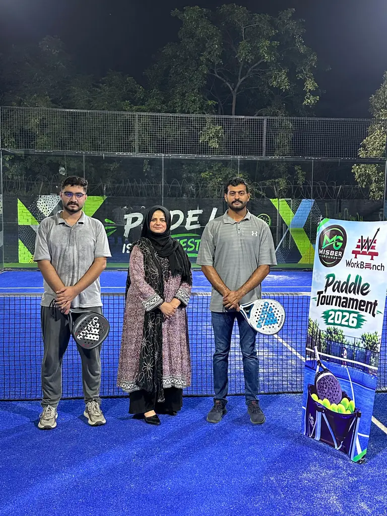 Two men holding padel rackets standing either side of a woman on a blue padel court at night, beside the Paddle Tournament 2026 banner and a court wall painted with the word padel.
