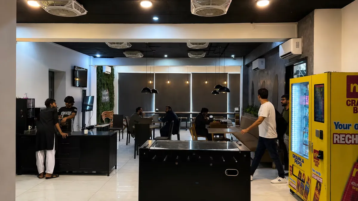 The cafeteria floor at WorkBench: coffee machines and a wall-mounted screen on the left, members seated at dining tables under dome pendant lights, a foosball table in the foreground and yellow vending machines on the right.