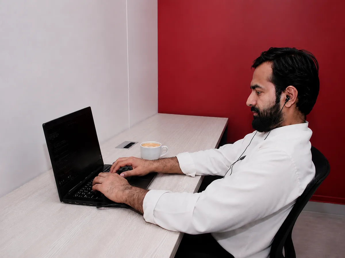 A member with earphones in, working on a laptop at a desk against a red accent wall at WorkBench, a coffee beside the keyboard.
