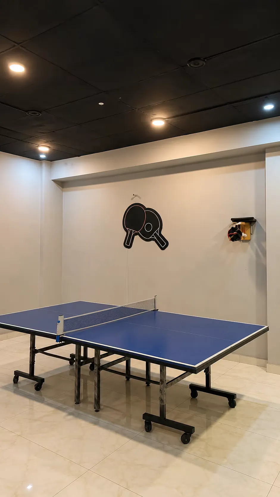 The game room at WorkBench: a blue table tennis table on a tiled floor under a black ceiling, with a painted paddle logo and a bat rack mounted on the wall.