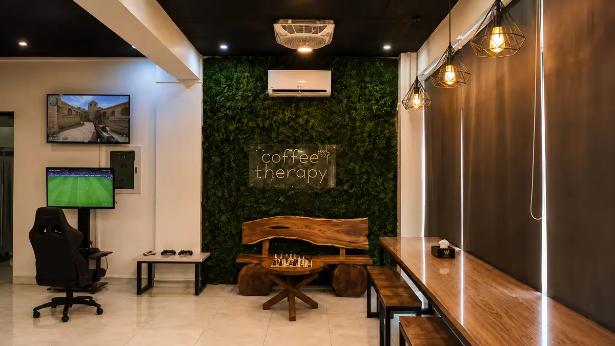 The gaming corner at WorkBench: a racing-style gaming chair facing two wall-mounted screens with controllers on a bench beside it, the coffee therapy nook and its moss wall visible beyond.
