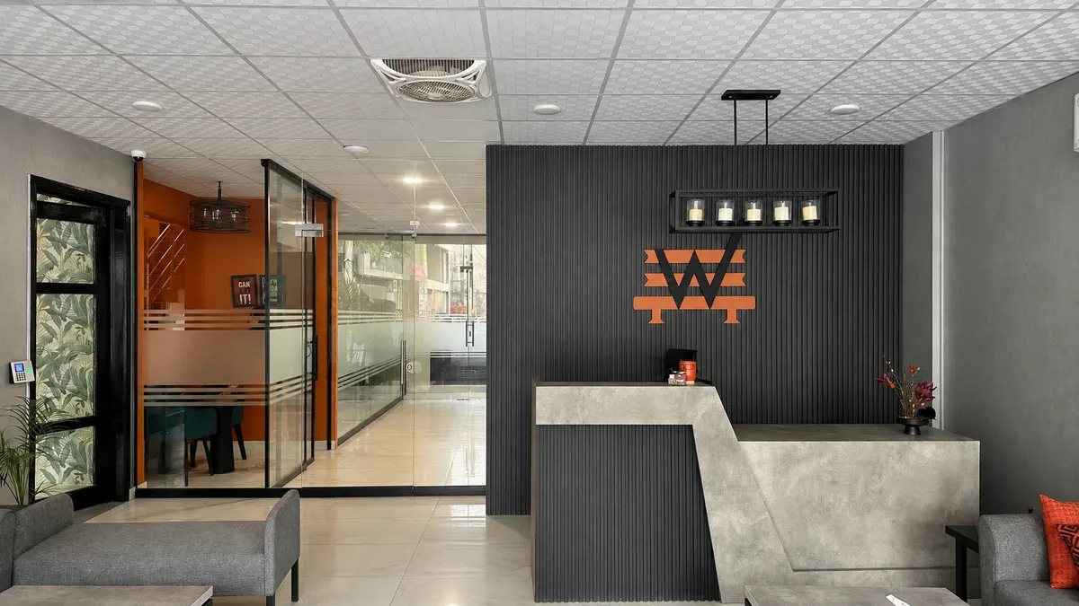 Reception at WorkBench: a concrete desk against a charcoal fluted wall with the amber W above it, a row of caged pendant lights, and a glass walled office to the left.