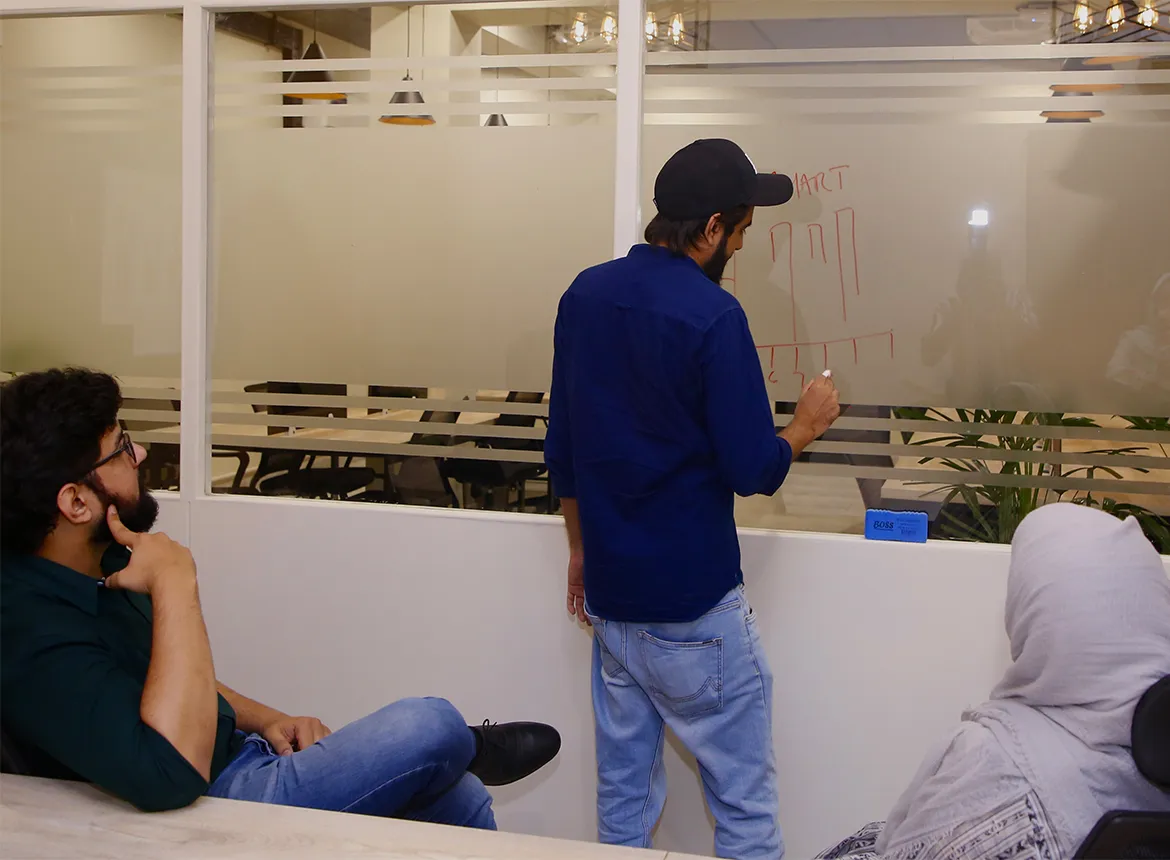 A man in a cap sketching a chart in red marker on a frosted glass partition at WorkBench while a colleague leans on the table to watch and another person in a grey headscarf sits in the foreground.